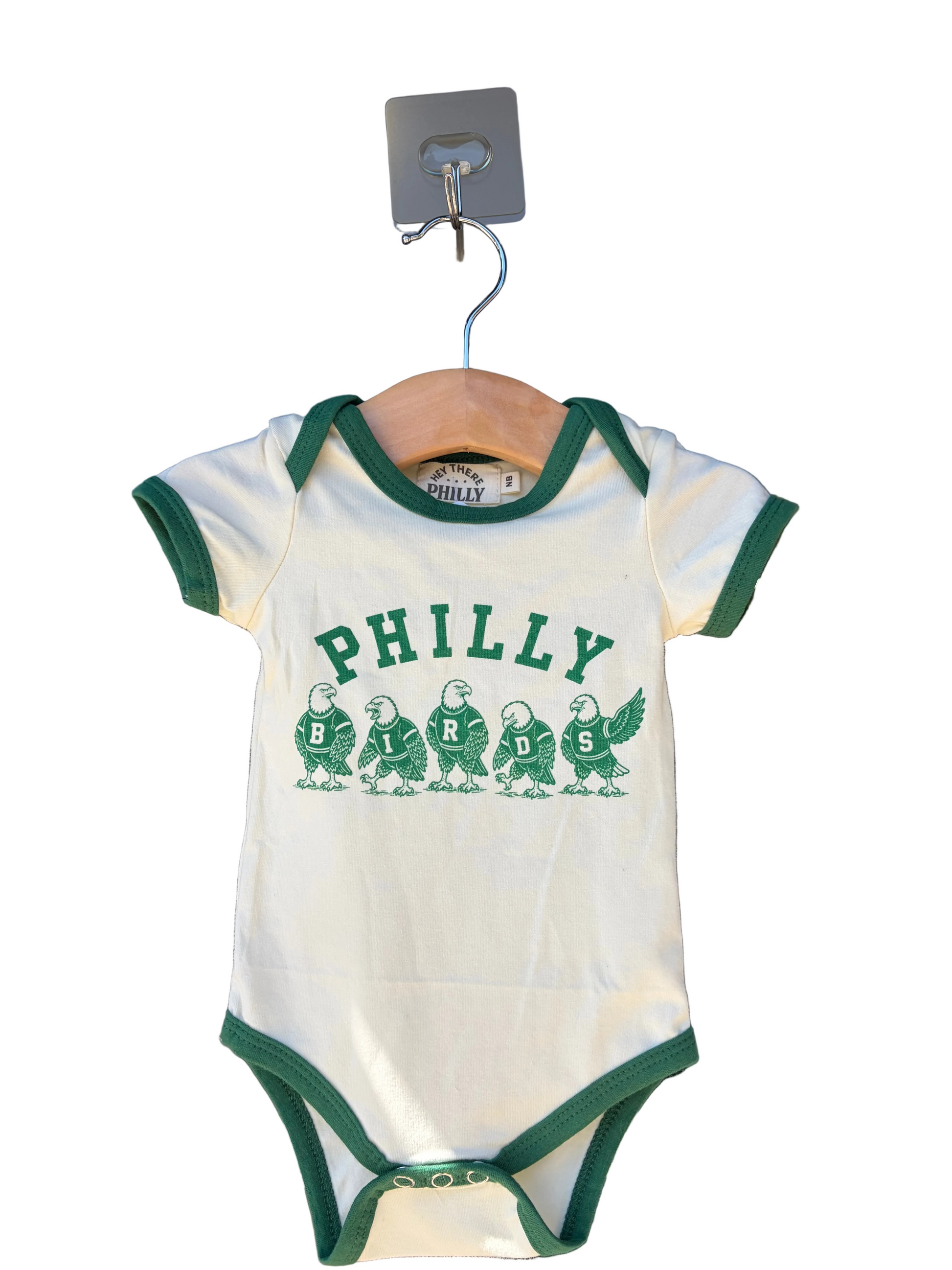 A white short-sleeve baby bodysuit with green trim and a green 'PHILLY' graphic featuring five partridge chicks, hanging on a wooden hanger in front of a glass storefront window.
