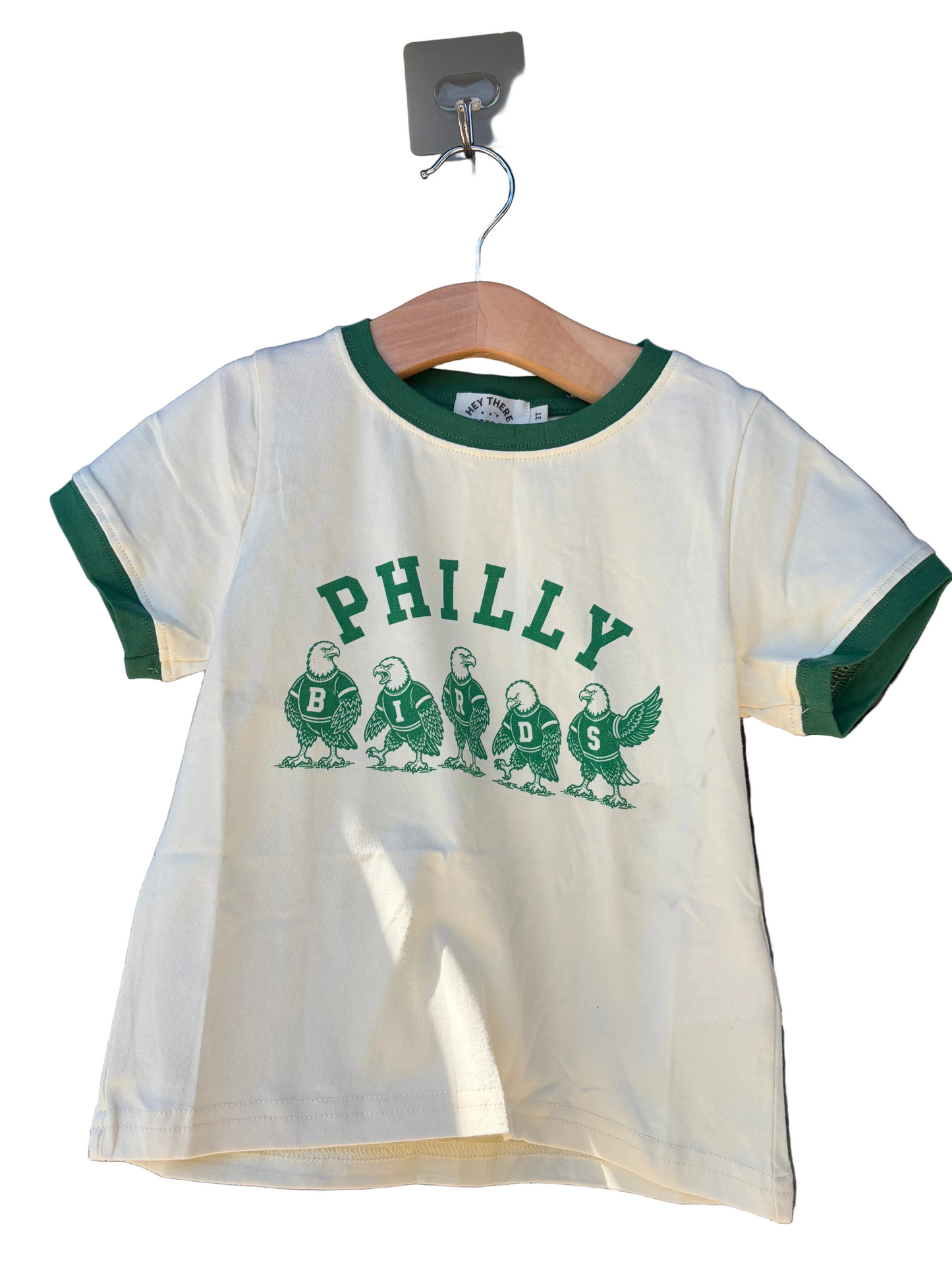 An off-white ringer t-shirt with green trim on the collar and cuffs, featuring a green graphic of five Philadelphia Eagles and the word 'PHILLY' across the chest, hanging on a wooden hanger.